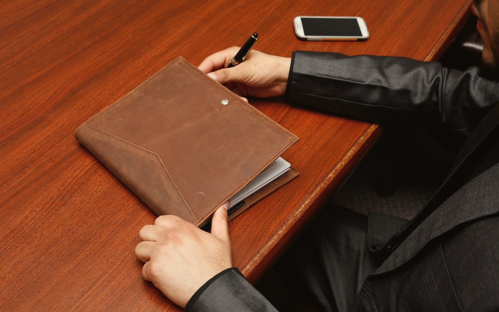 A person in a suit writes with a pen on a leather portfolio at a wooden desk
