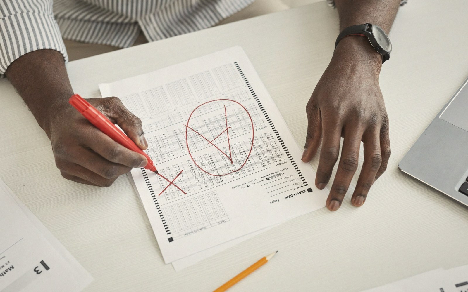 Hands marking checkmarks and an x on a multiple-choice exam form with a red pen
