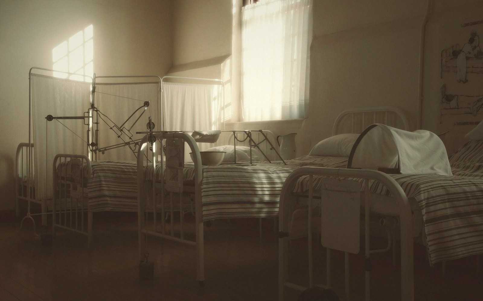 Old hospital ward with metal-frame beds, striped linens and sunlight through curtained windows