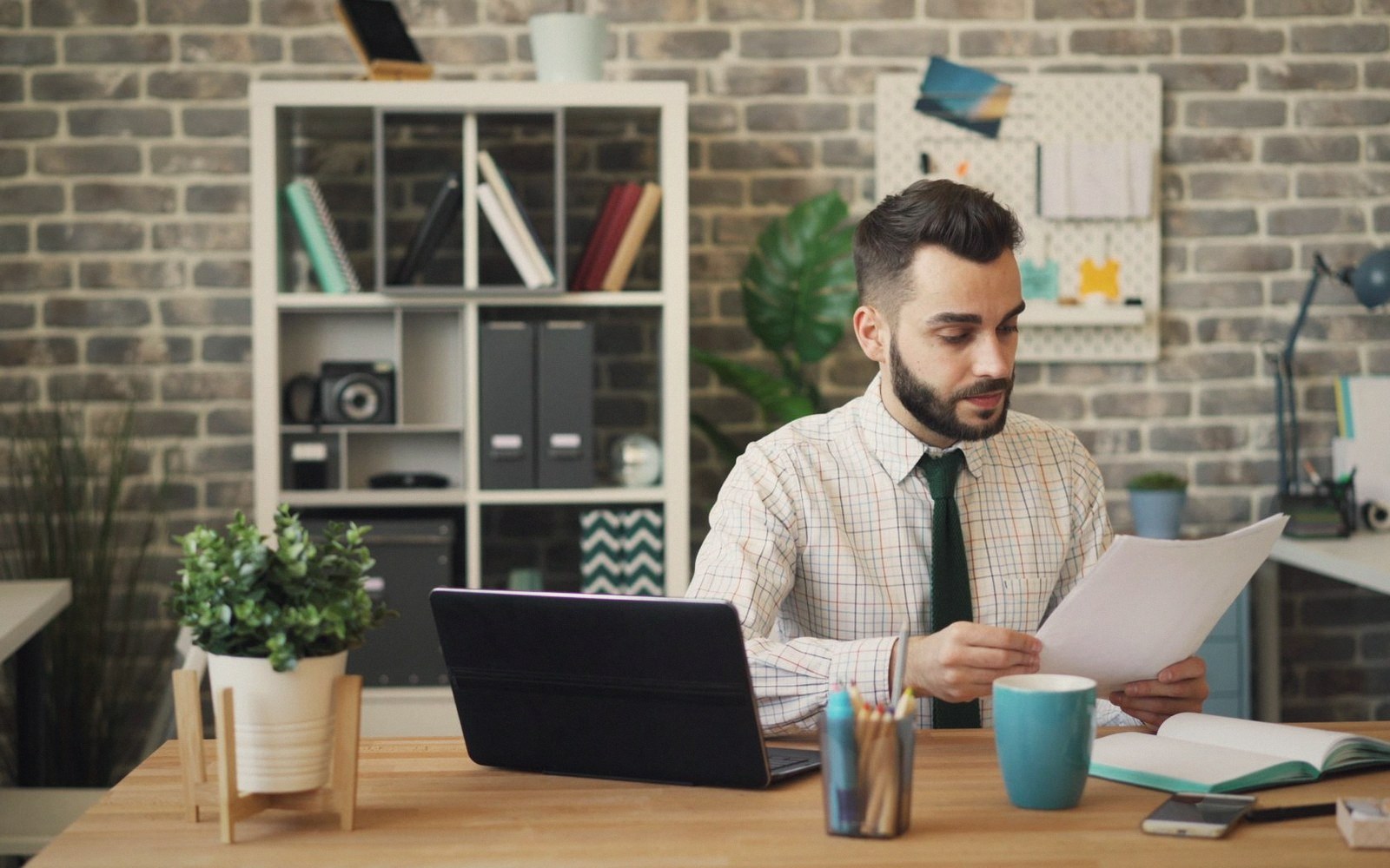 Man in shirt and tie reviews papers at a desk with a laptop and coffee mug