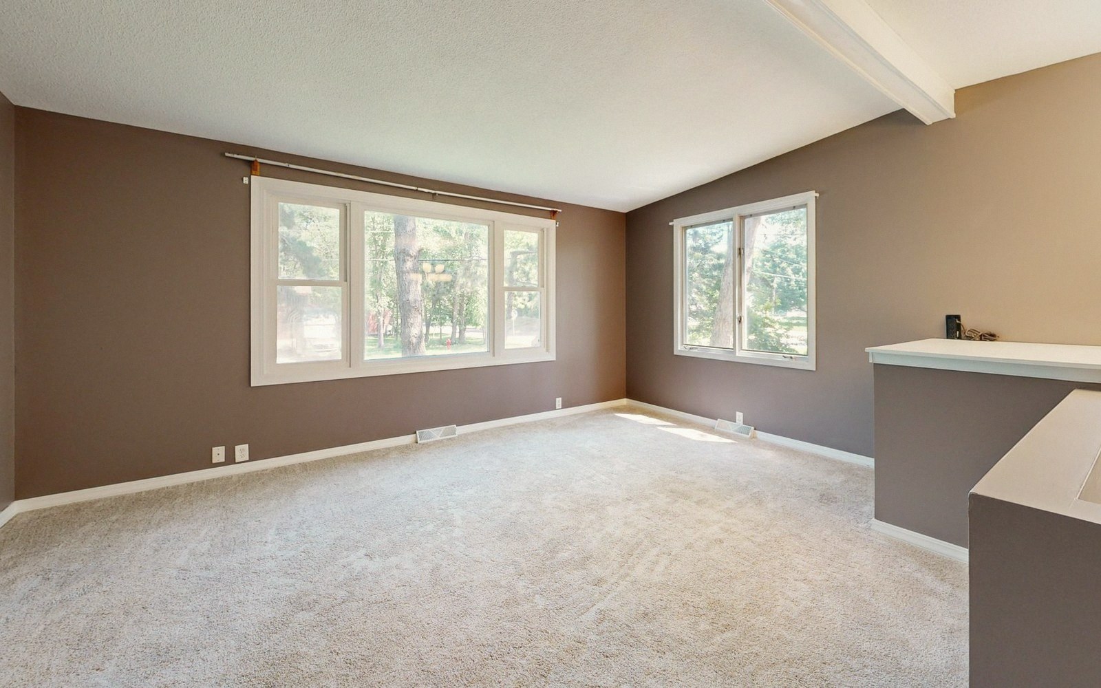 Empty carpeted living room with two windows and a breakfast bar counter visible on the right