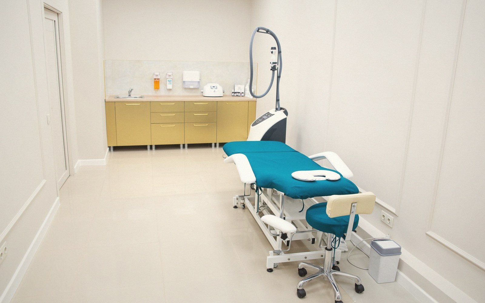 A treatment room with a teal examination chair, stool, overhead device, and cabinetry